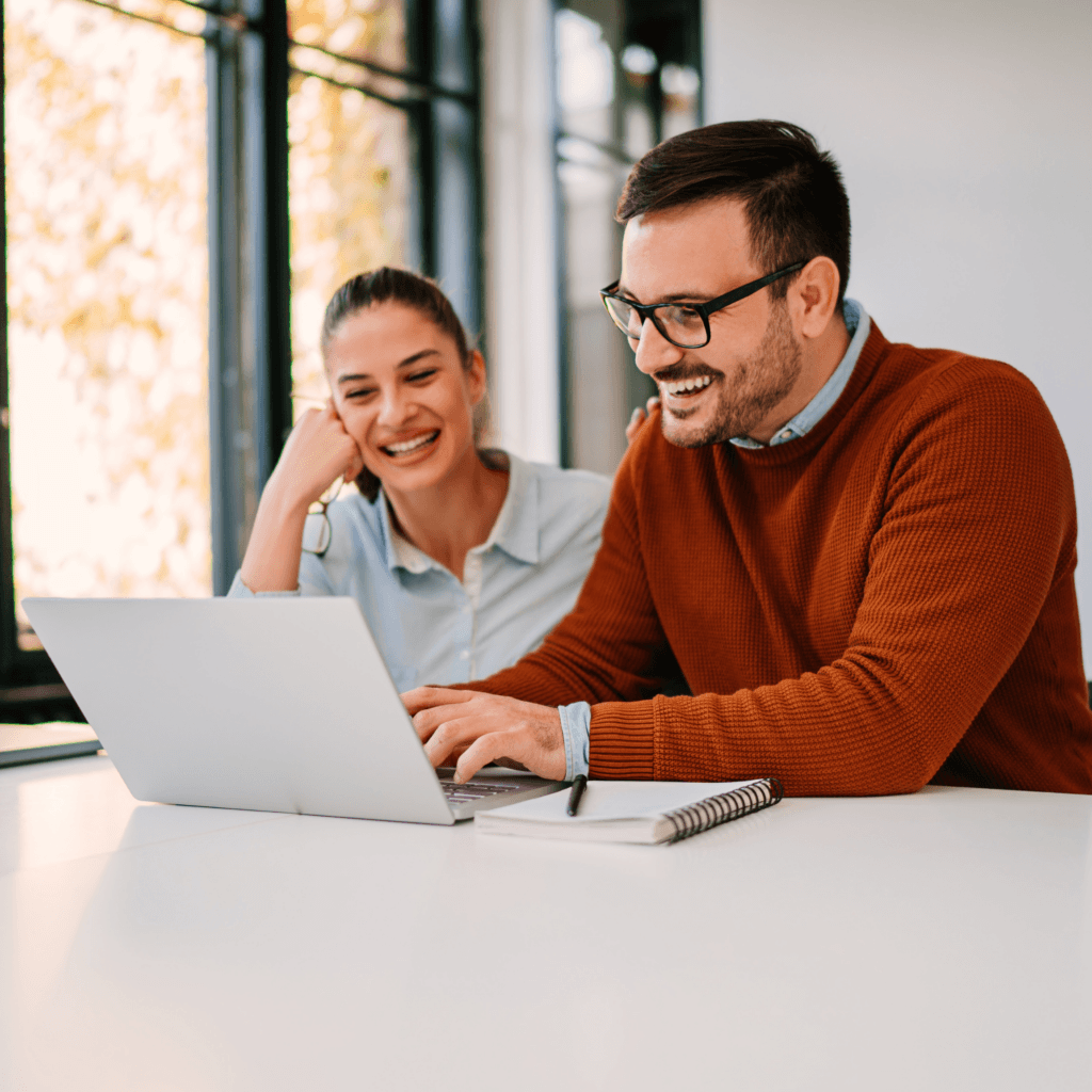 Two background screening company employees sat at an office desk smiling at a laptop.