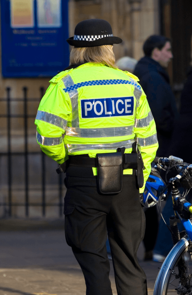A UK police officer in a bright yellow jacket and hat stands near a bicycle on a city street.