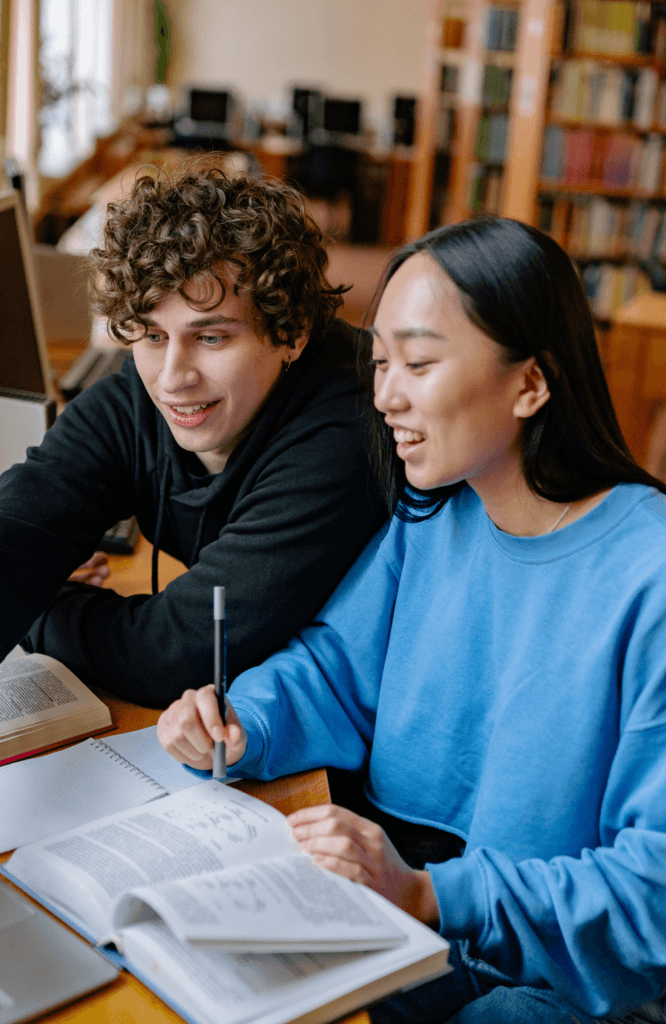Two students studying together at a library table, surrounded by open books and focused on their work.