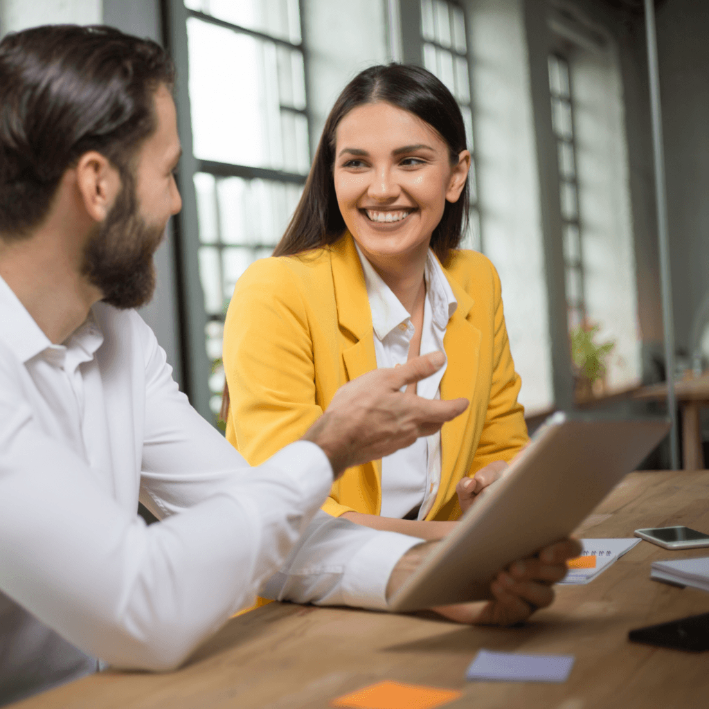 Two colleagues are happily discussing work at a desk in a well-lit office, with documents and a tablet in view.