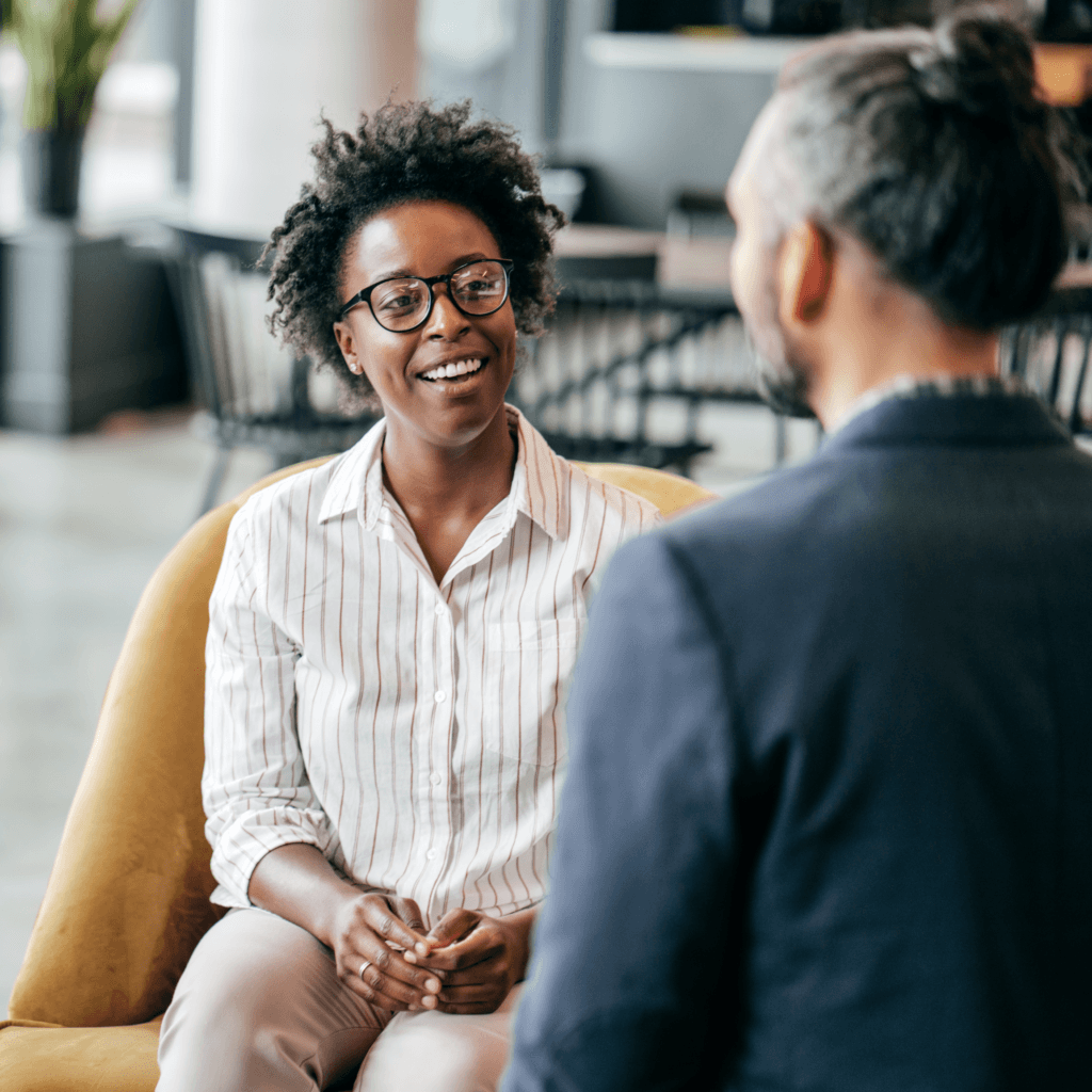 Two people are conversing in a modern office setting, one seated on a mustard chair, smiling and engaged, representing the team of a background screening services organisation.