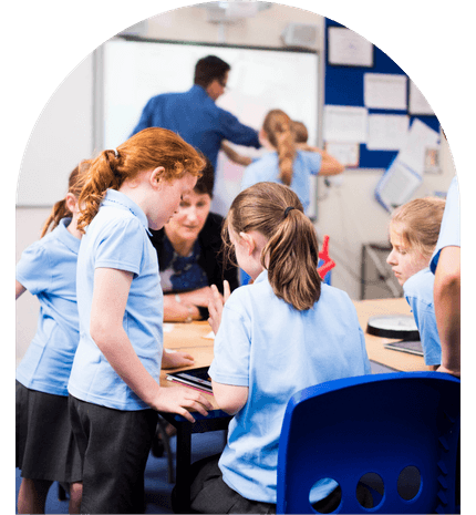 Children in a classroom engaged in group work, using tablets, with a teacher assisting in the background.