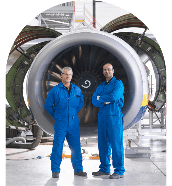 Two engineers in blue coveralls stand confidently in front of a large jet engine in a hangar.