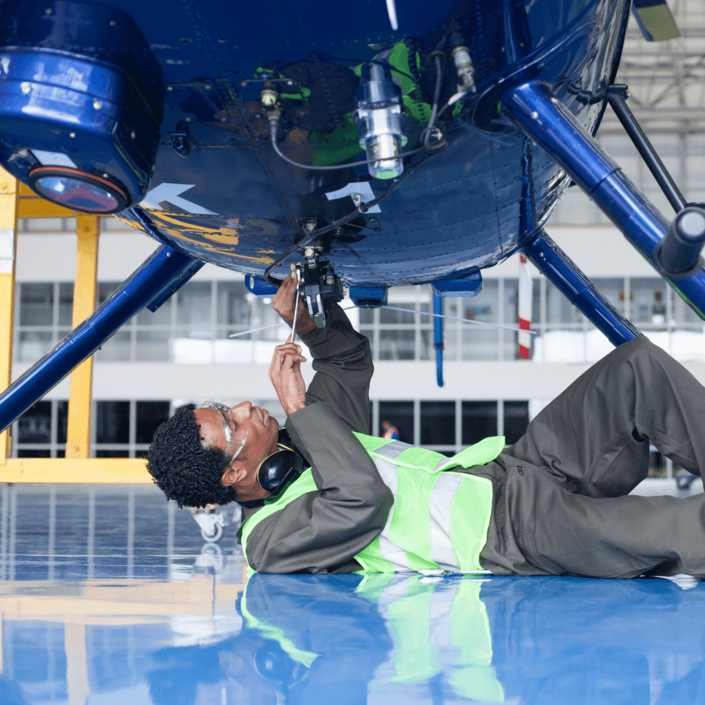 aerospace and defence background screening - Engineer lying under helicopter, performing maintenance in a brightly lit hangar.