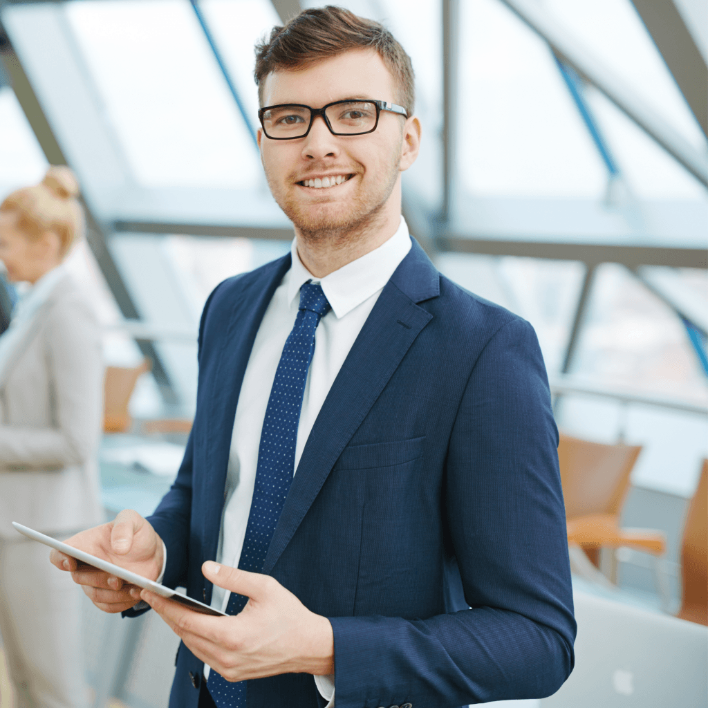 A professional services employee, a young man, in a suit and glasses smiles while holding a tablet in a modern office setting.