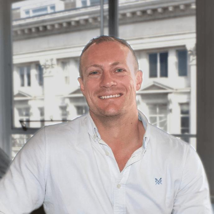 Smiling person in a white shirt sits in front of a window with an urban building background.