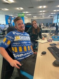 Two people smiling at a desk in an office, with one wearing a festive jumper.