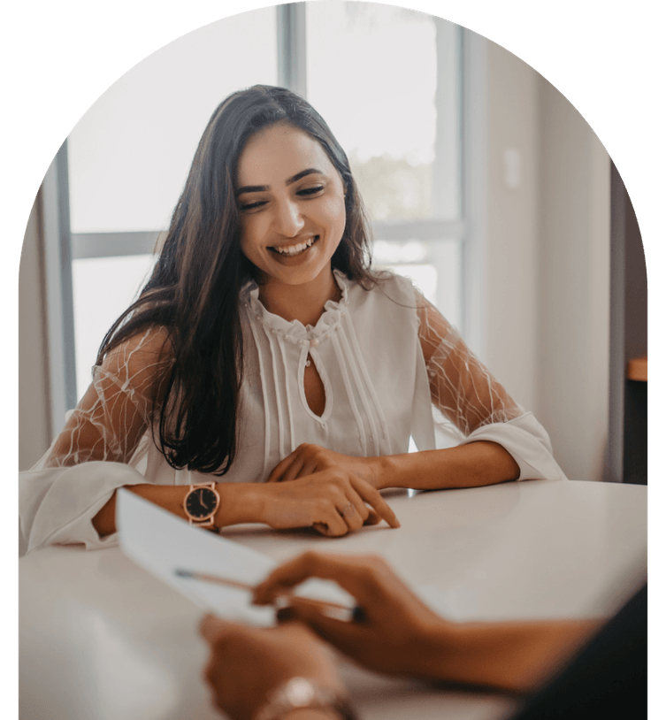 A young woman in a white blouse sits at a table, smiling, during a meeting, with a person opposite holding a document, representing a successful interview after use of talent pool.