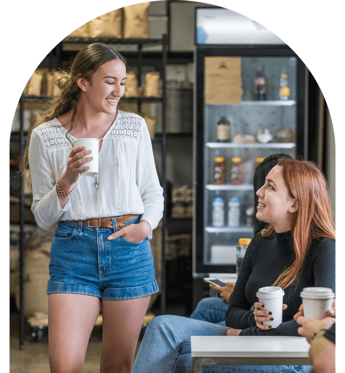 Two young adults smiling and chatting over coffee in a cosy café setting, with a fridge and shelves in the background.