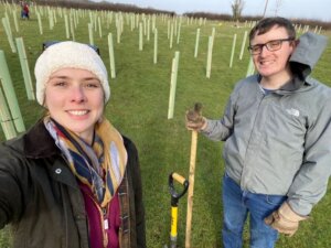 Two people smiling in a field with young tree saplings, holding a spade, on a cloudy day.