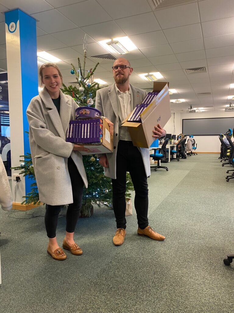 Two people in an office carrying gift boxes stand by a decorated Christmas tree.