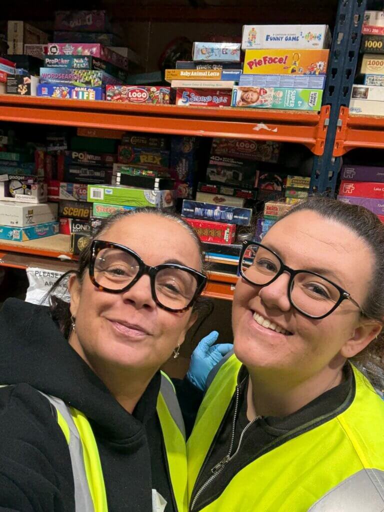 Two people wearing glasses and high-visibility vests smiling in front of shelves filled with board games.