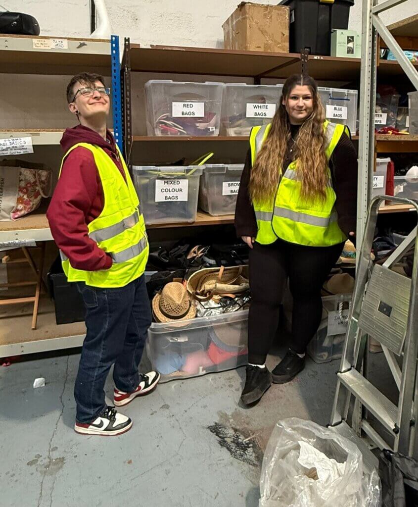 Two people wearing high-visibility vests stand in a warehouse, surrounded by labelled boxes and shelves.