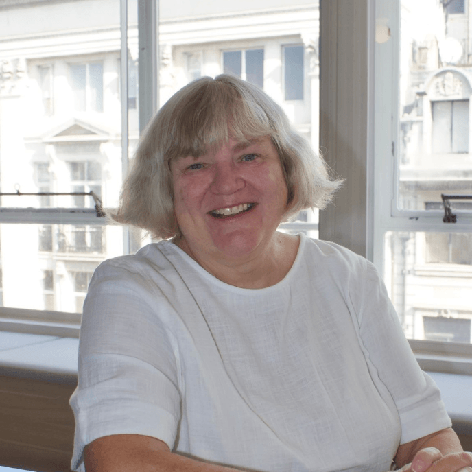Workforce Management Experts - Leslie Weare smiling with short blonde hair sits by a window, wearing a white shirt, with city buildings in the background.