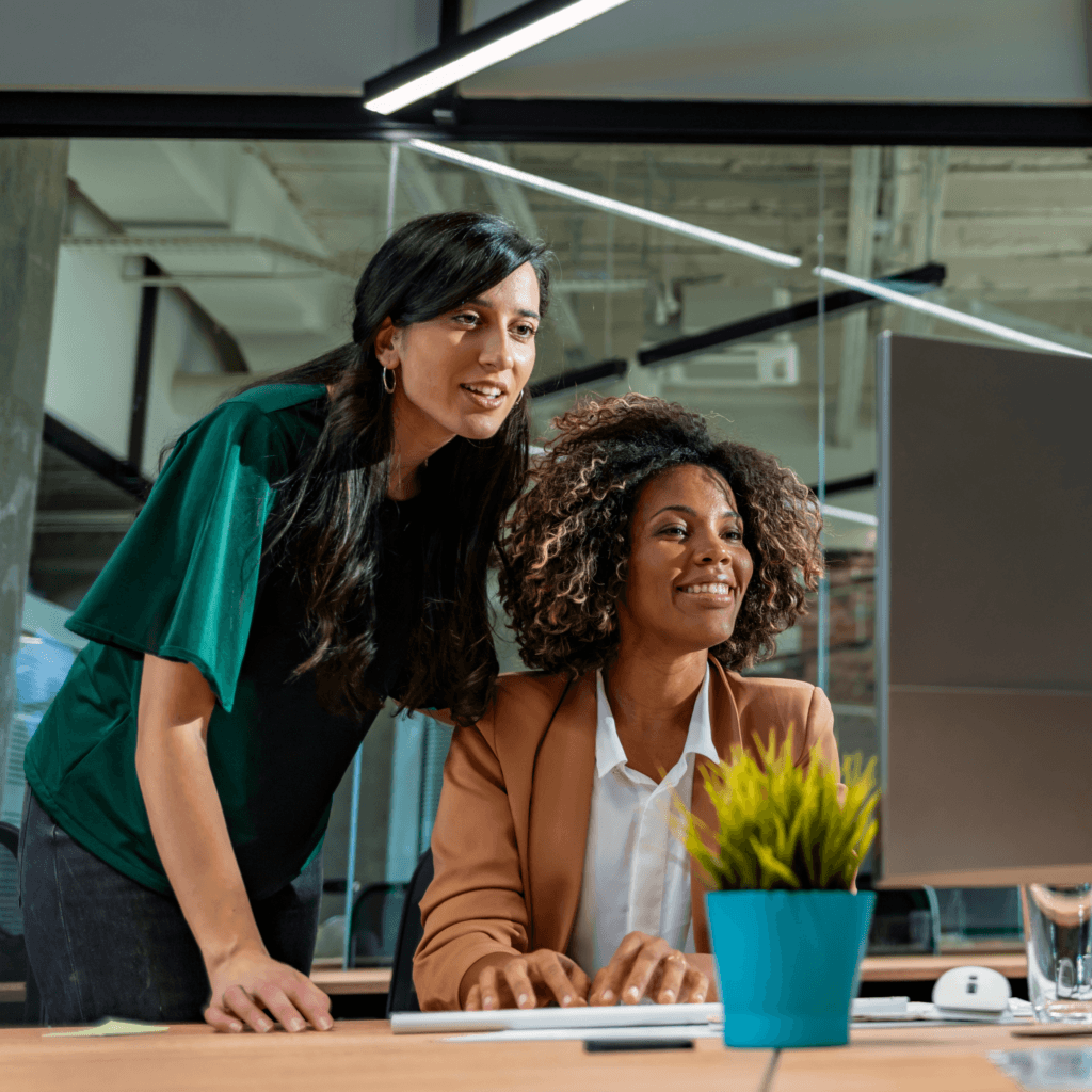 Two colleagues smiling while working at a computer in a modern office, with a small potted plant in the foreground, representing Umbrella Solutions team.