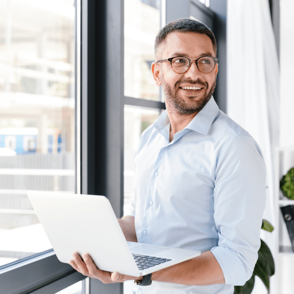 A man in glasses and a light blue shirt, smiling while holding a laptop near a window, looking care free and engaged with his work - signalling the ease and benefit of Total Workforce Management from Matrix.