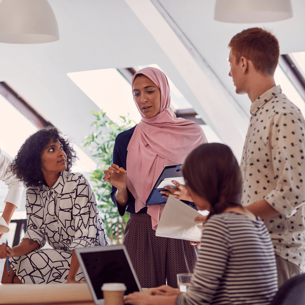 A diverse group of colleagues engaged in a discussion around a table in a modern, well-lit office space.