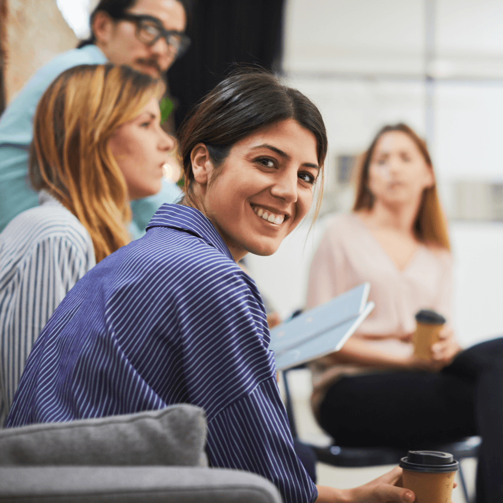A group of four people in a casual meeting setting, with one smiling woman in focus holding a cup, representing workforce management solutions team.