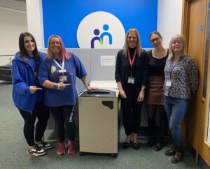 Five women standing together in an office setting, smiling at the camera in front of a blue and white logo.