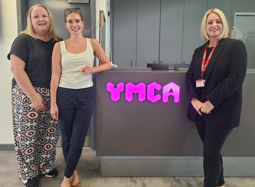 Three people smiling in front of a YMCA reception desk with a pink illuminated sign.