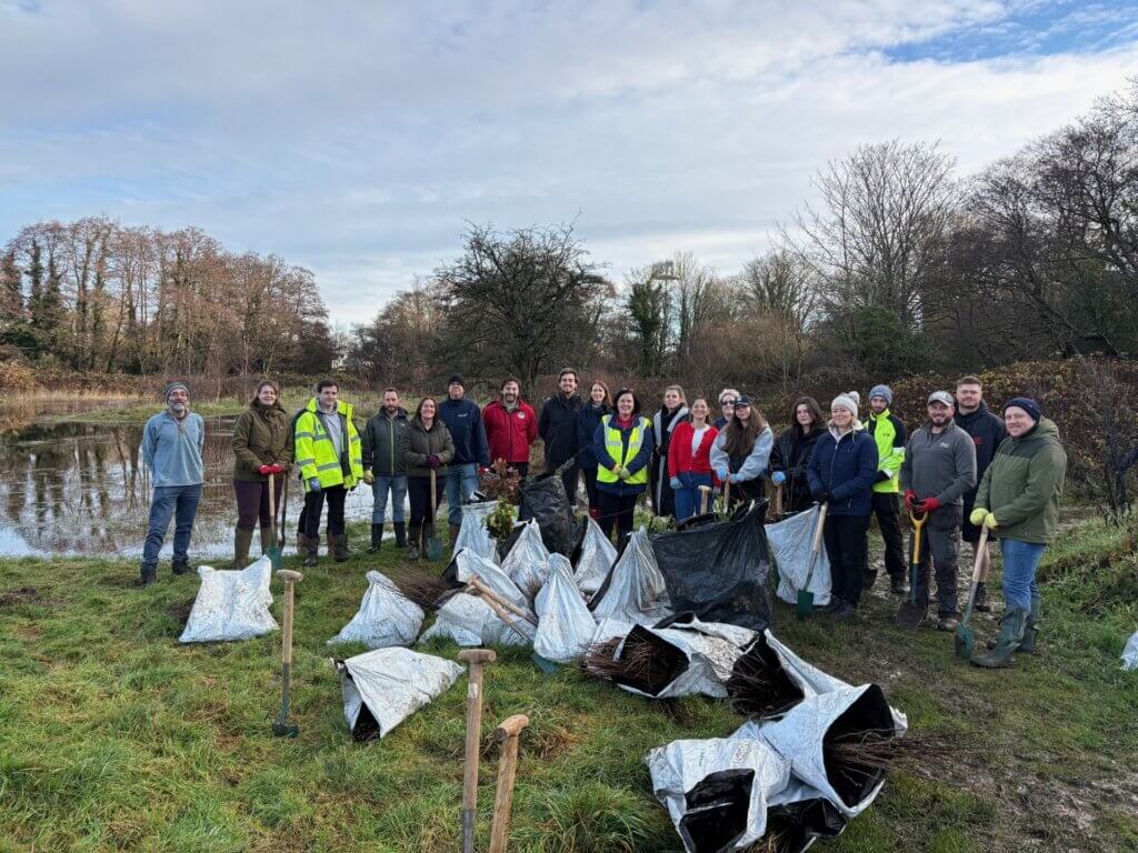 Group of people gathered outdoors near a pond, with piles of filled bags and tools on the grass.