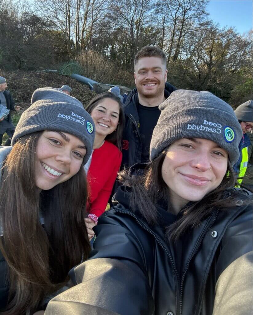 A group of smiling people outdoors in winter clothing, wearing matching grey beanies, with trees in the background.
