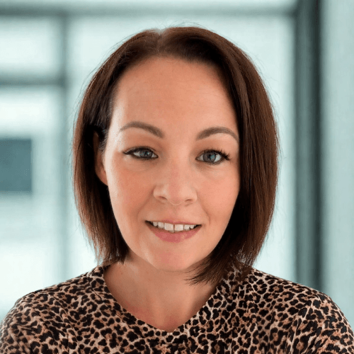 A person with short brown hair wearing a leopard print top, smiling slightly in a light-filled room.