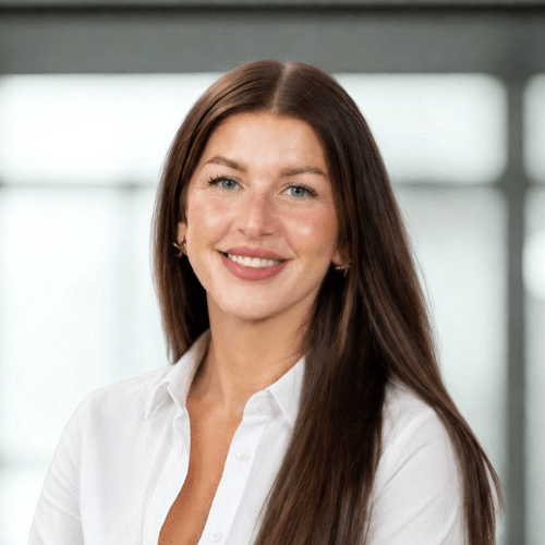 A smiling person with long brown hair wearing a white shirt in front of a blurred background.